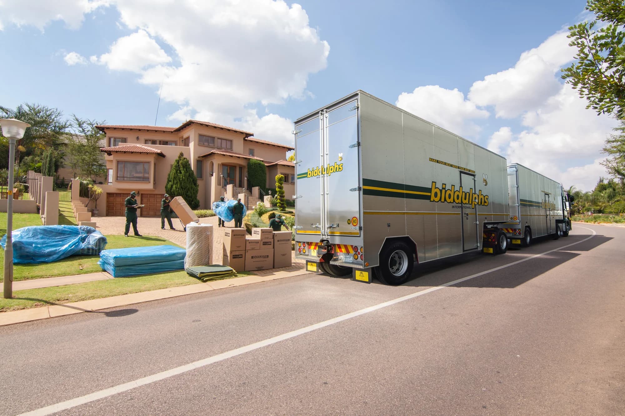 A Biddulphs truck and trailers beside cartons and wrapped furniture outside a home